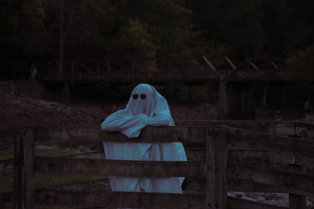 Image of a person dressed as a ghost, with sunglasses over the sheet and leaning on a fence